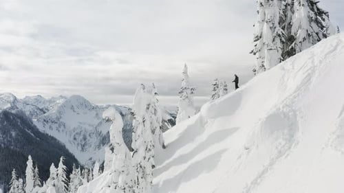 Snowboarder On Top Of Snowy Mountain Peak Looking Down The Ski Slope Aerial Drone Flying Past