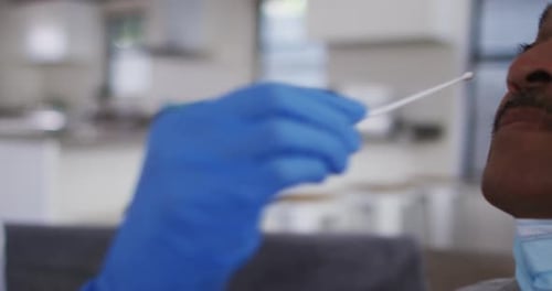 Mixed race female doctor wearing mask doing swab test on senior man at home