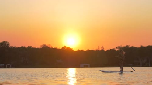A young woman sup stand-up paddleboarding on a lake at sunset.