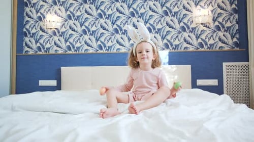 Une jolie petite fille aux cheveux longs est assise au centre d'un grand lit et joue avec un lapin décoratif