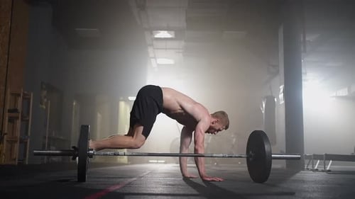 A Young Man Without a Shirt in Slow Motion Jumps a Burpee Over a Barbell in the Gym in Backlight