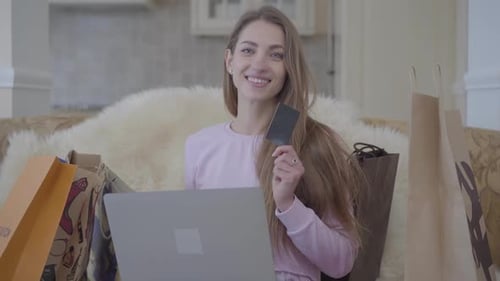Cute Woman Sitting on a Sofa with a Laptop and Lots of Shopping Bags Near, Showing a Black Credit
