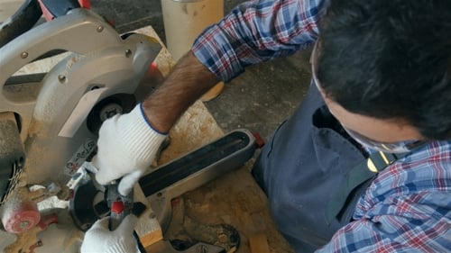 Carpenter Works With Wooden Bar In Workshop Near Electric Saw.