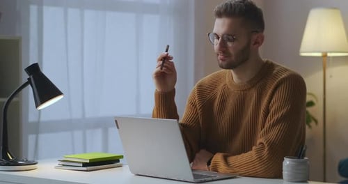 Thoughtful Man Is Sitting in Front of Laptop in Room and Thinking Spinning Pen in Hand Writer or