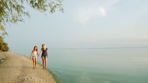 Mom And Daughter Walk Along The Picturesque Beach Where The Blue Sky Merges With The Water
