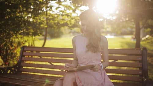 Young Woman Sitting On The Bench In a Park And Reading a Book. Beautiful European Girl Studying