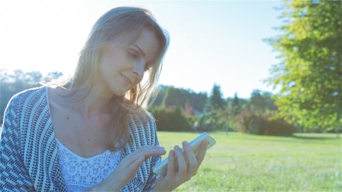 Happy Smiling Girl Using a SmartPhone in City Park Sitting on Grass