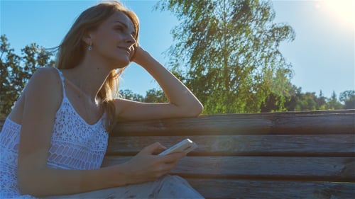 Happy Smiling Girl Using SmartPhone in City Park Sitting on the Bench