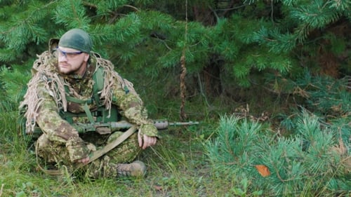 The Young Man In Uniform With a Weapon In His Hands. He Is Sitting In The Forest, Resting, Looking