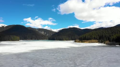Aerial view of a frozen mountain lake surrounded by forest and snowy mountains in spring season.