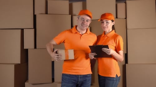 Couriers In Orange Uniform Standing Against Brown Carton Stacks Backround. Delivery Company Staff