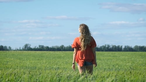 Beautiful Blonde Young Romantic Woman In a Red Shirt Walking Slowly At The Green Field
