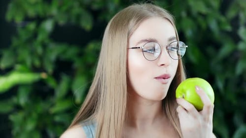 Young Woman Eats Green Apple in Front of Plants