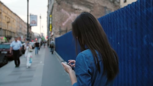 Young Woman Stands Near a Blue Fence In The Street. Lady Uses a Smartphone, Turns Her Head To The