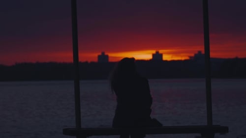 Silhouette eines Mädchens, das auf einer Bank sitzt und den Sonnenuntergang bewundert.