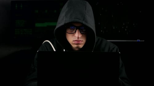 Hacker With Glasses And In a Hooded Working On a Computer In a Dark Office Room