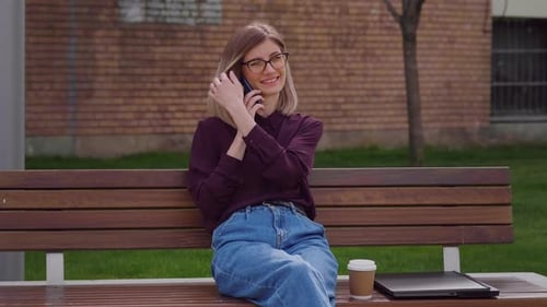 Business woman talking and laughing with her mobile phone while sitting on a bench in park.