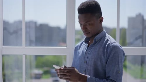 Young Focused Man Typing on Smartphone Screen Standing at Window in Home Office