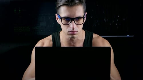 Male Hacker With Glasses Working On a Computer In a Dark Office Room