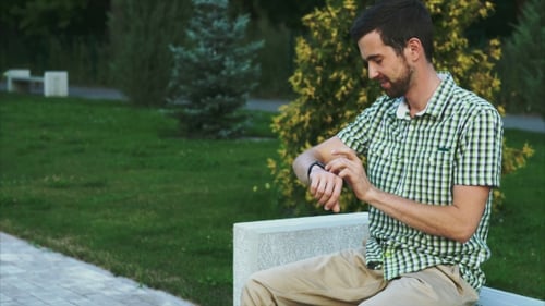 Casual Dressed Man Sit On The Bench In The Park And Use His Modern Smart Watch