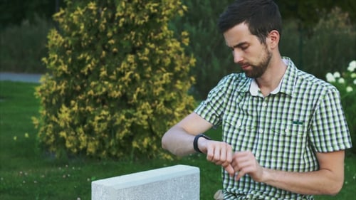 Young Man Using Smartwatch While Sitting On The Bench In The Park