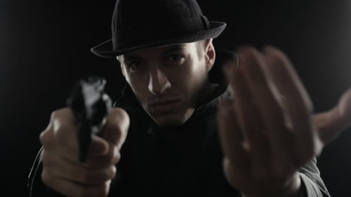 Gangster In a Hat And a Black Cloak. Mafioso Aiming a Gun At The Camera