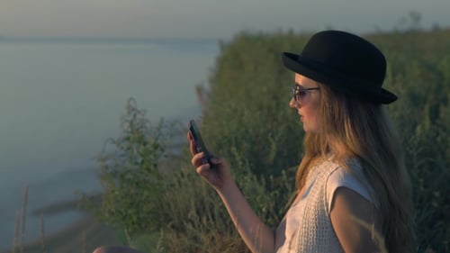 Young Girl Use Her Smartphone Sitting On The Edge Of a Cliff Near The River