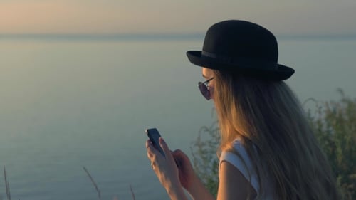 Young Girl Use Her Smartphone Sitting On The Edge Of a Cliff Near The River