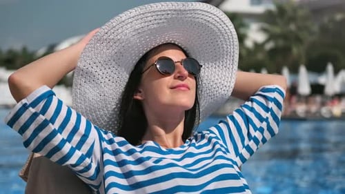Femme souriante décontractée avec lunettes de soleil et chapeau blanc prenant un bain de soleil près de la piscine, gros plan