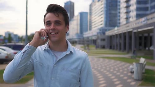 A successful young man in casual clothes is walking along a city street and talking on the phone.