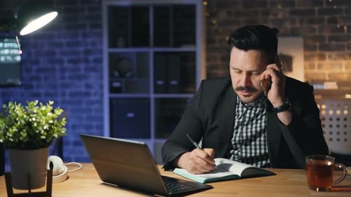 Serious Man Office Worker Talking on Mobile Phone and Using Laptop at Night