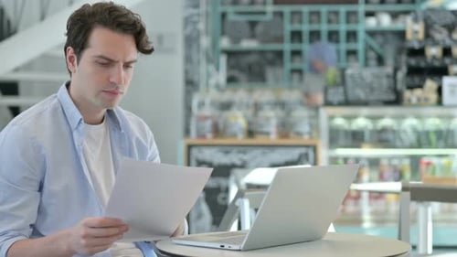 Man Reading Papers, Reacting Negatively at Laptop