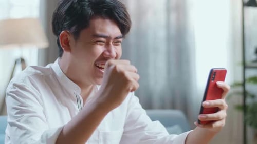 Close Up Side View Of Happy Asian Man Celebrating While Using Smartphone In The Living Room