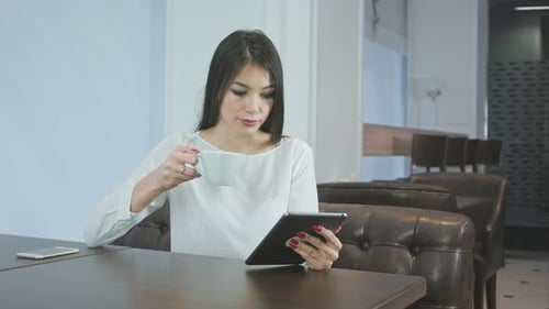 Young Woman Using Digital Tablet While Drinking Coffee or Tea at a Cafe