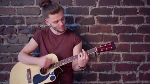 Young Adult Plays Acoustic Guitar against Brick Wall