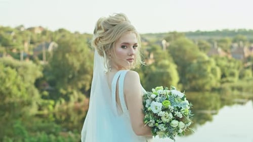 Happy Young Woman Bride with a Wedding Bouquet Stands on the Background of the River