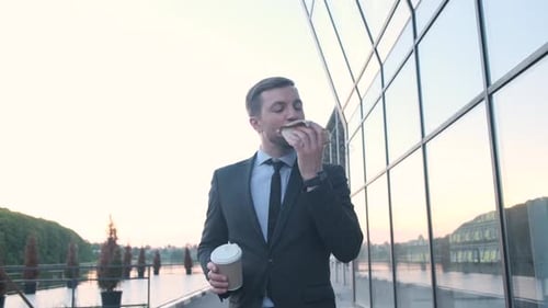 Young Handsome Businessman Holding a Cup of Coffee Eating a Burger on the Street