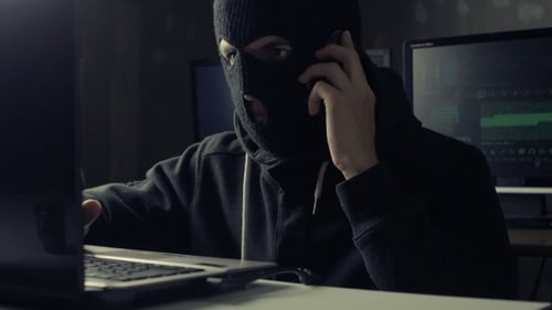 Dangerous Hacker In a Mask Speaks Phone At a Computer In a Dark Office Room