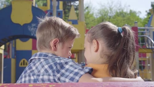 Netter blonder Junge und ein hübsches Mädchen sitzen auf der Bank vor dem Spielplatz und umarmen sich. Ein Paar