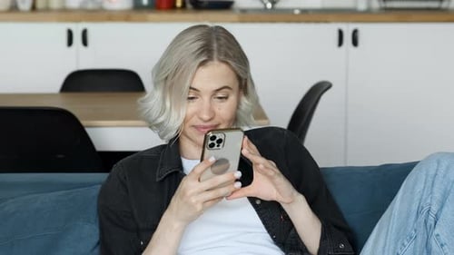 Young Caucasian attractive blonde woman looks at the screen, texting a message on the smartphone