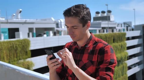 Attractive Man in Plaid Shirt Using Smartphone Touching Screen Outdoors at Urban City Background