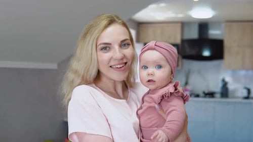 Mother Holds Baby in Her Arms on the Background of a Modern Kitchen Set and Looks Into the Camera