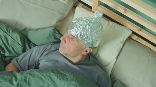 Un hombre con una gorra de aluminio yace en la cama sosteniendo un letrero de cartón que dice «FALSO»