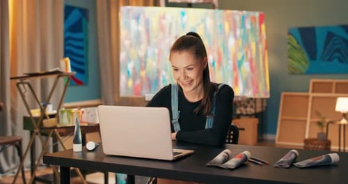 Artist Dressed in Work Clothes Sitting in Studio Behind Painting on Canvas Standing on Easel