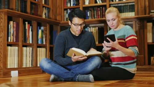 Friends Sitting on the Floor in the Library. A Man Holding an Open Book, a Woman Used a Tablet