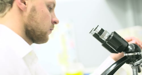 Scientist Is Looking Through Microscope and Writing Data on Tablet.