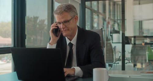 A businessman talking on the phone while working on a laptop at the office