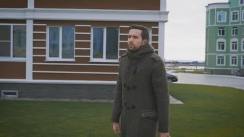 A Man in a Gray Coat in the Autumn Weather Walks on a Footpath. Against the Background of the City