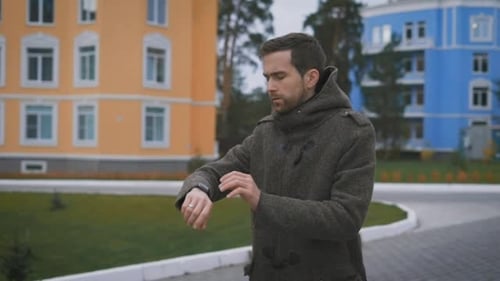 of Young Bearded Man in Brown Coat Walking in the Street Near Buildings and Checking His Watch