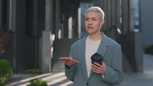 Woman with Coffee Walks and Uses Phone Outdoors